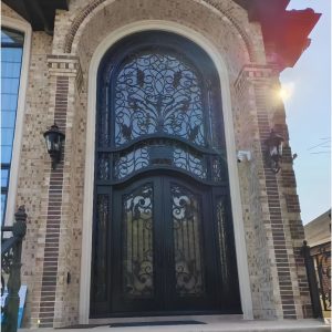 A Premium Cast Iron Spiral Staircase by Like Metal features intricate scrollwork and a black finish, showcased in an elegant interior setting with sunlight streaming in from above and decorative wall lanterns nearby.