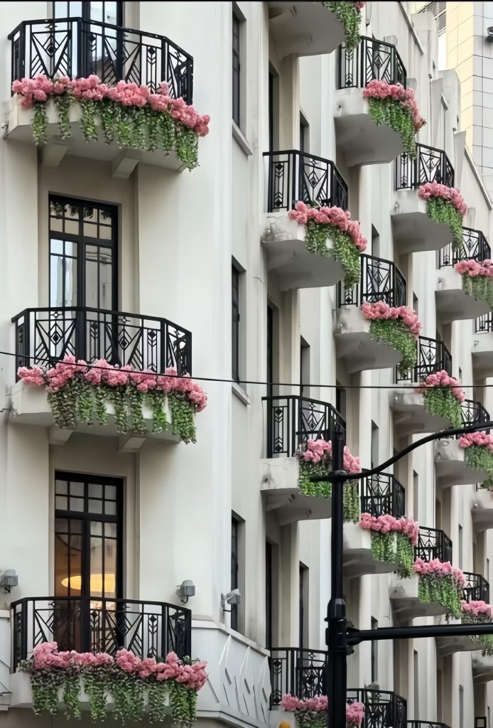 A row of balconies with ornate black railings, featuring Premium Cast Iron Spiral Staircases by Like Metal, each decorated with pink flowers and trailing green plants on a white building.