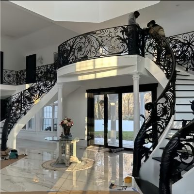A grand foyer with a marble floor, a round table with flowers, large glass doors, and a dramatic Premium Cast Iron Spiral Staircase by Like Metal featuring ornate railings. Two people are working near the top of the stairs.